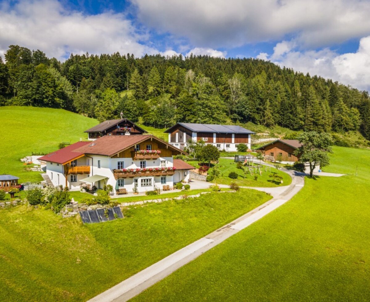 The Grubsteighof Farm House, situated on green hills backed by a forest, features multiple buildings and a children's playground.