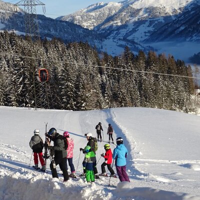 Skiers on a snow-covered mountain slope with a ski lift, surrounded by evergreen forests and distant peaks.