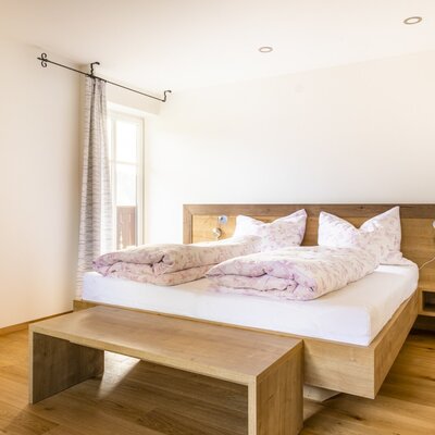 The Farm House bedroom features a wooden double bed with integrated bedside shelves, reading lights, and power outlets, a wardrobe, a wooden bench, and natural light from the window.
