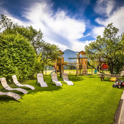 The Farm House garden features a lawn with sun loungers, a children's play area including a playground and basketball hoop, and mountain views.