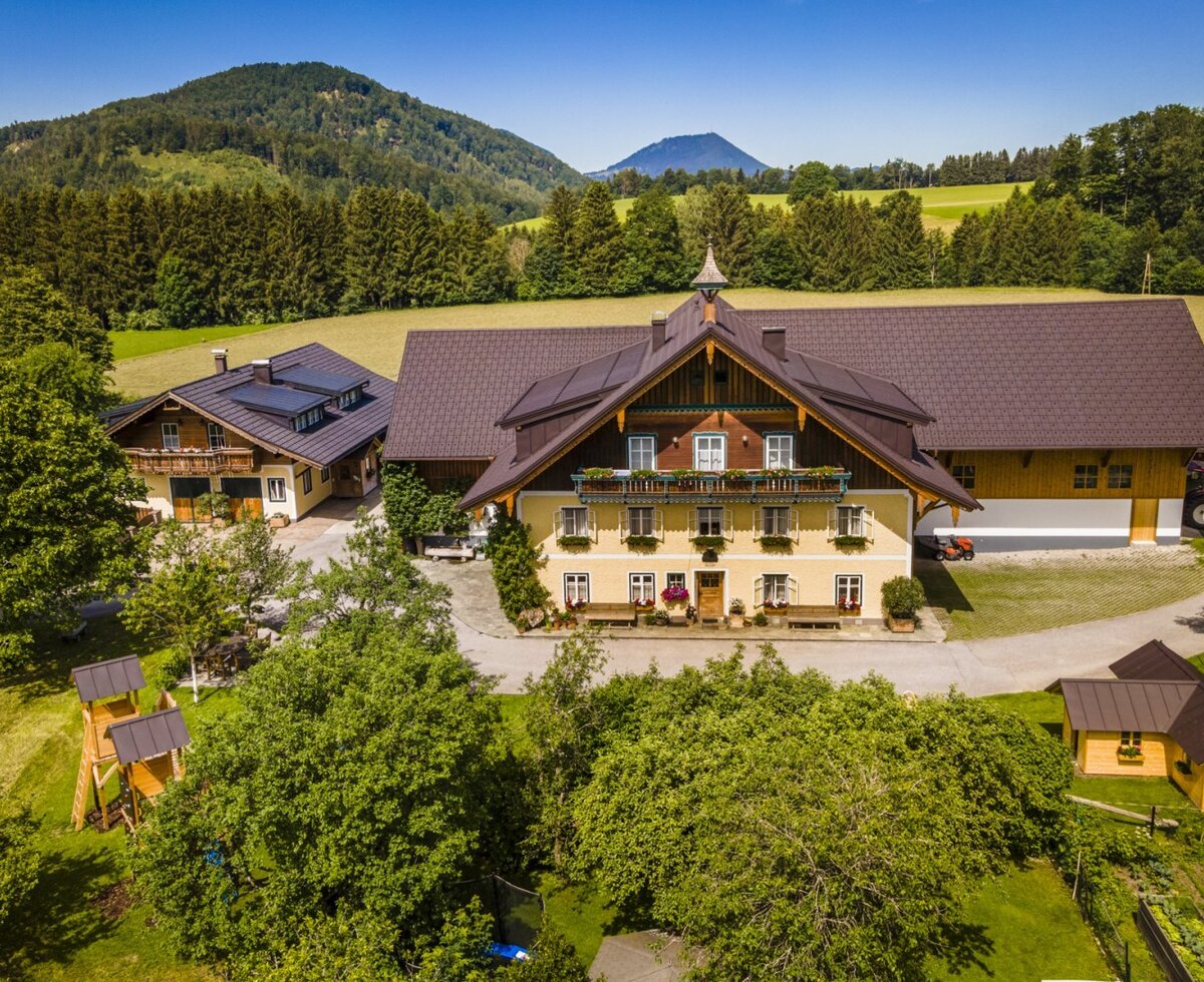 The farm house, featuring multiple buildings, a garden with a children's play structure, and surrounding mountain and forest landscapes.