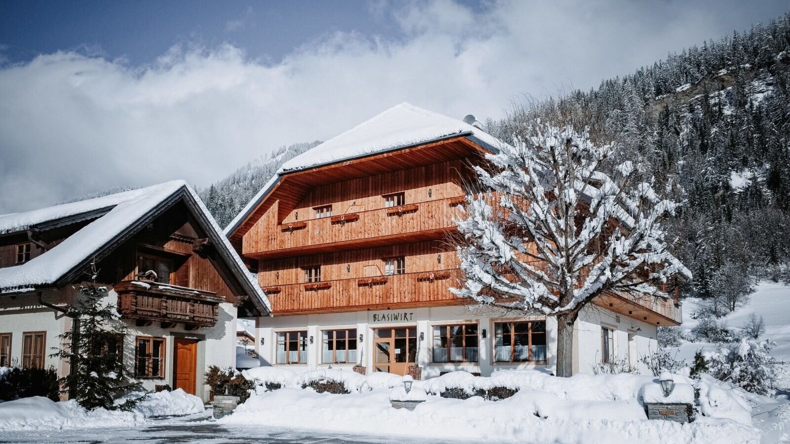 Exterior view of the farmhouse in winter, featuring snow on the roof, balconies, and surrounding landscape.