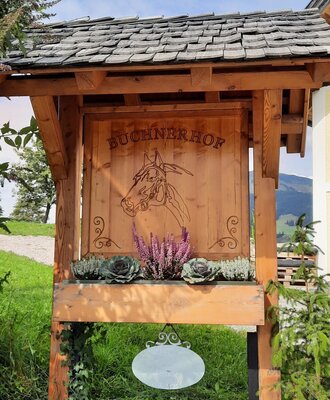 The wooden entrance sign for Buchnerhof Farm House, featuring a carved horse design and a planter with seasonal foliage.