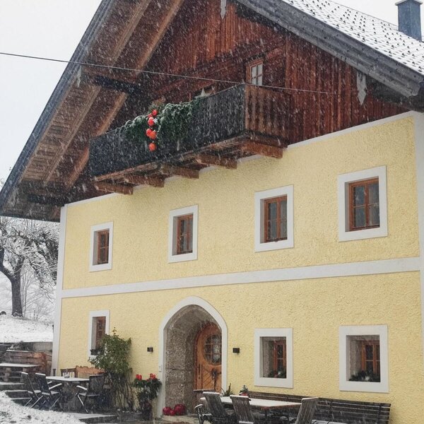 The exterior of the farmhouse, featuring a yellow facade, a wooden upper floor with a balcony, and outdoor seating during snowfall.