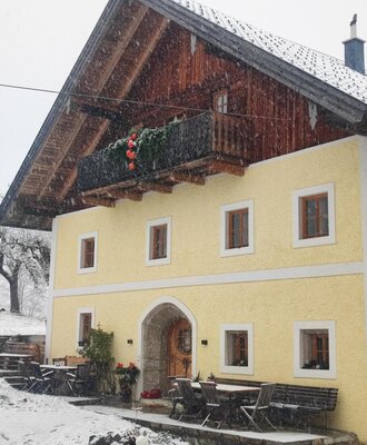 The exterior of the farmhouse, featuring a yellow facade, a wooden upper floor with a balcony, and outdoor seating during snowfall.