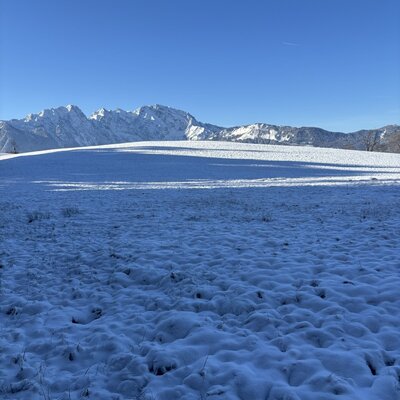 Snow-covered landscape with mountains in the background under a clear blue sky, surrounding the farmhouse.