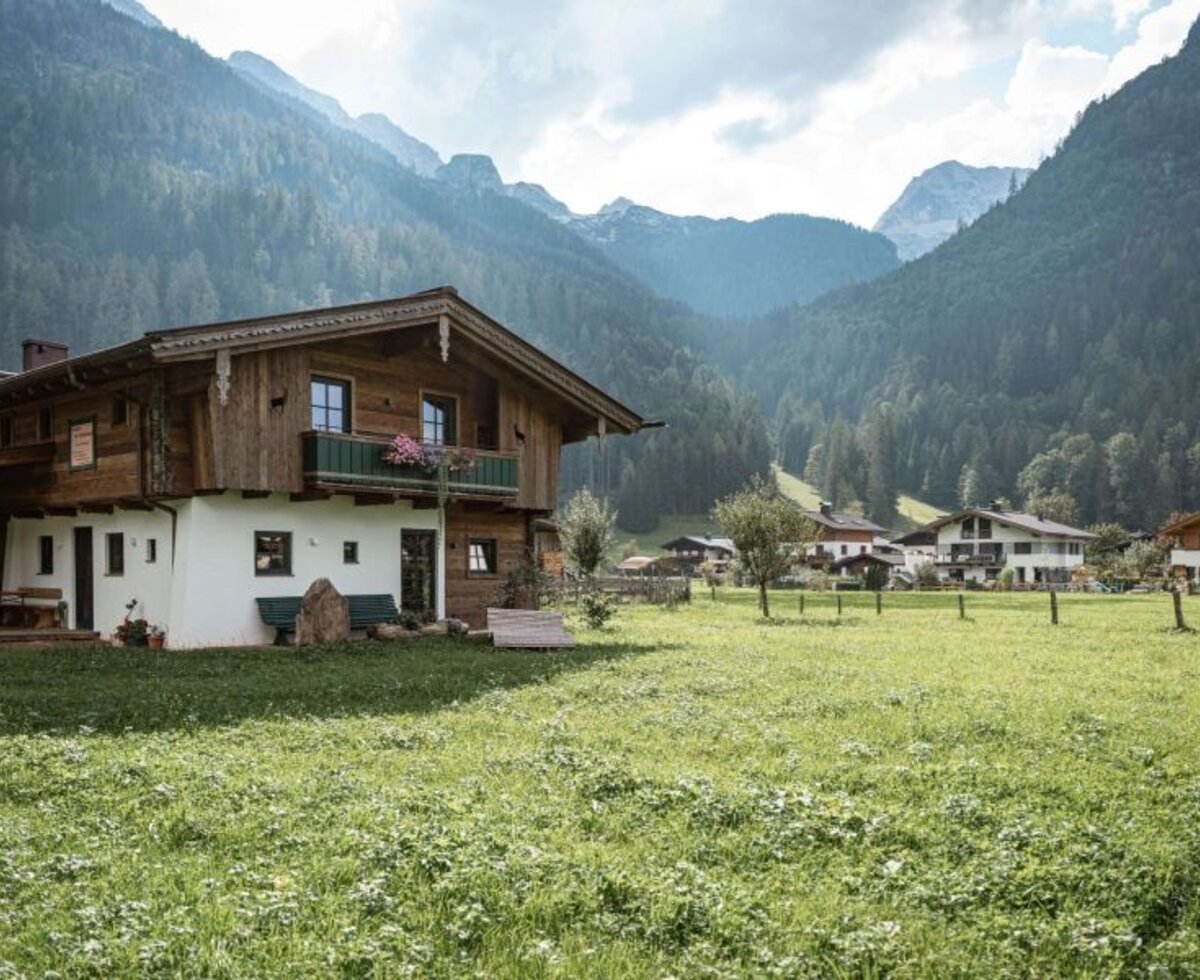 Exterior view of the Bed and Breakfast with traditional wooden facade, balcony, and surrounding meadow against a mountain backdrop.