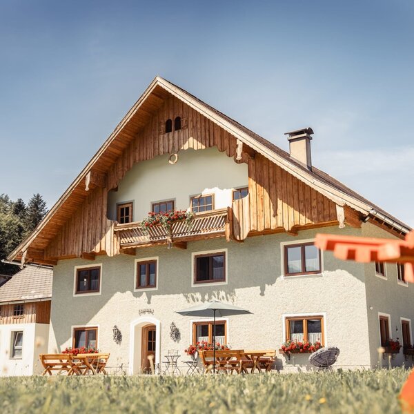 The Farm House exterior, featuring traditional wooden balconies with flower boxes, an outdoor terrace with tables and chairs, and a sun lounger on the surrounding lawn.