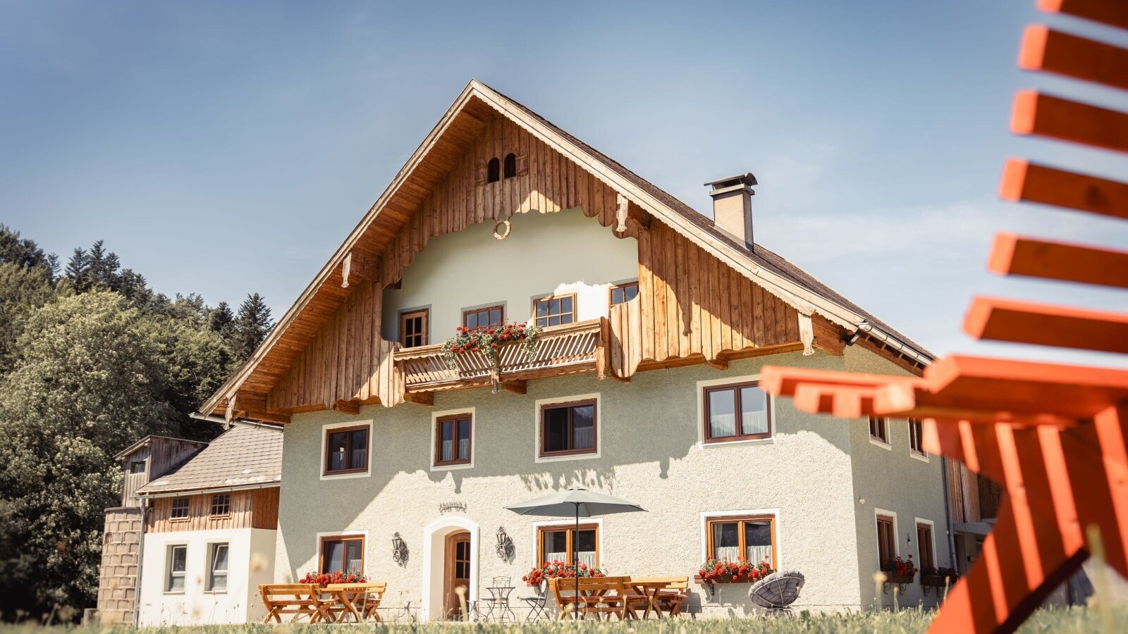 The Farm House exterior, featuring traditional wooden balconies with flower boxes, an outdoor terrace with tables and chairs, and a sun lounger on the surrounding lawn.