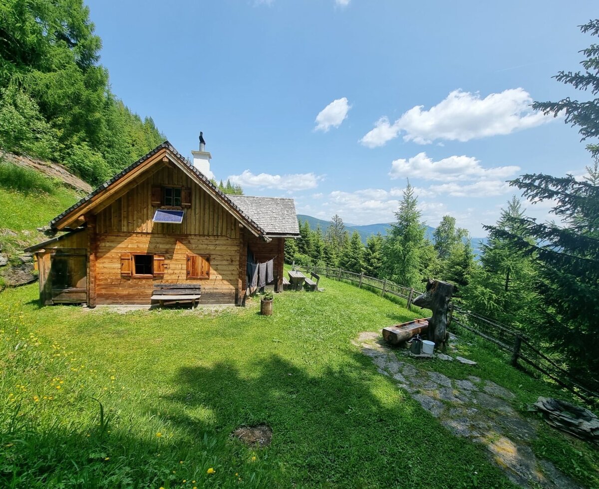 The Almhütte with a solar panel and outdoor seating, surrounded by meadows and mountains.
