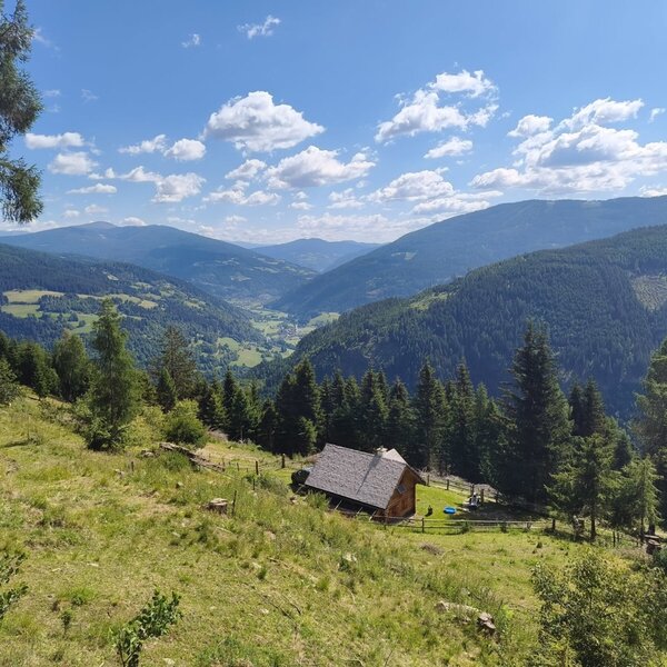 The rustic mountain hut nestled on a grassy hillside, offering scenic views of the surrounding valley and forested mountains.