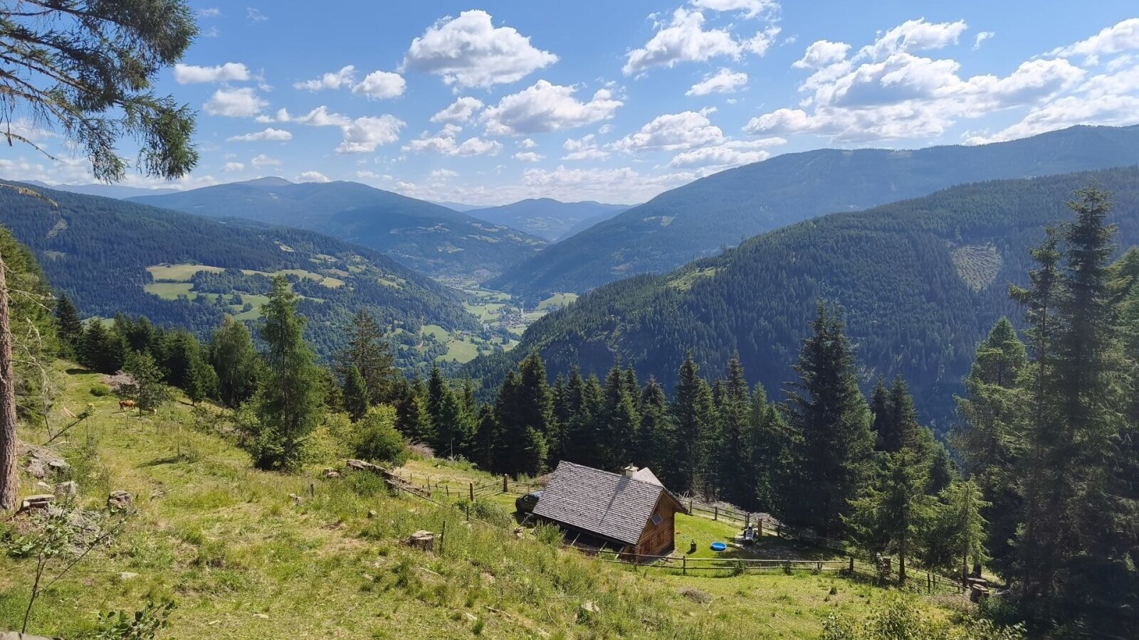 The rustic mountain hut nestled on a grassy hillside, offering scenic views of the surrounding valley and forested mountains.