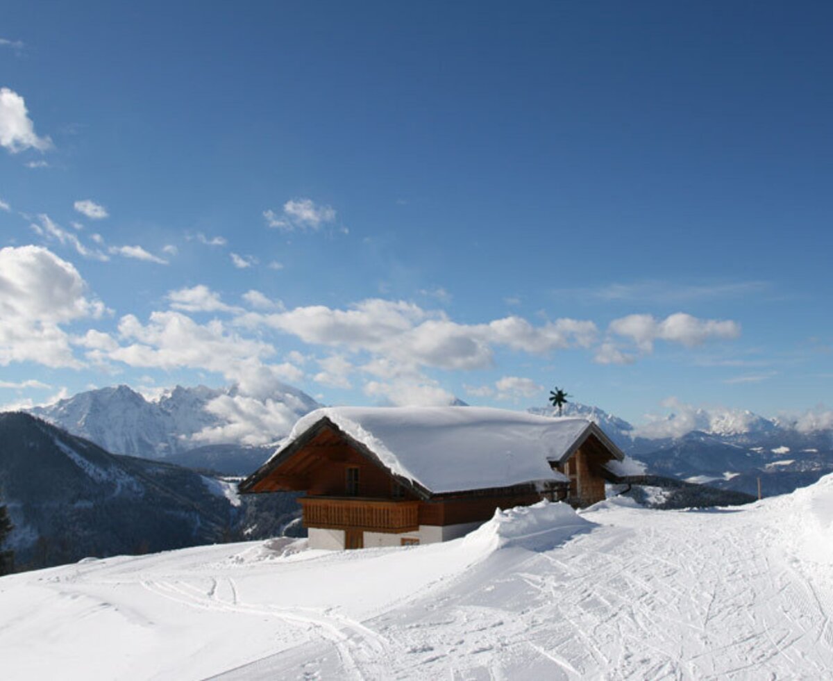 The snow-covered farmhouse is set on a mountain slope, with ski tracks in the foreground and distant snowy peaks.