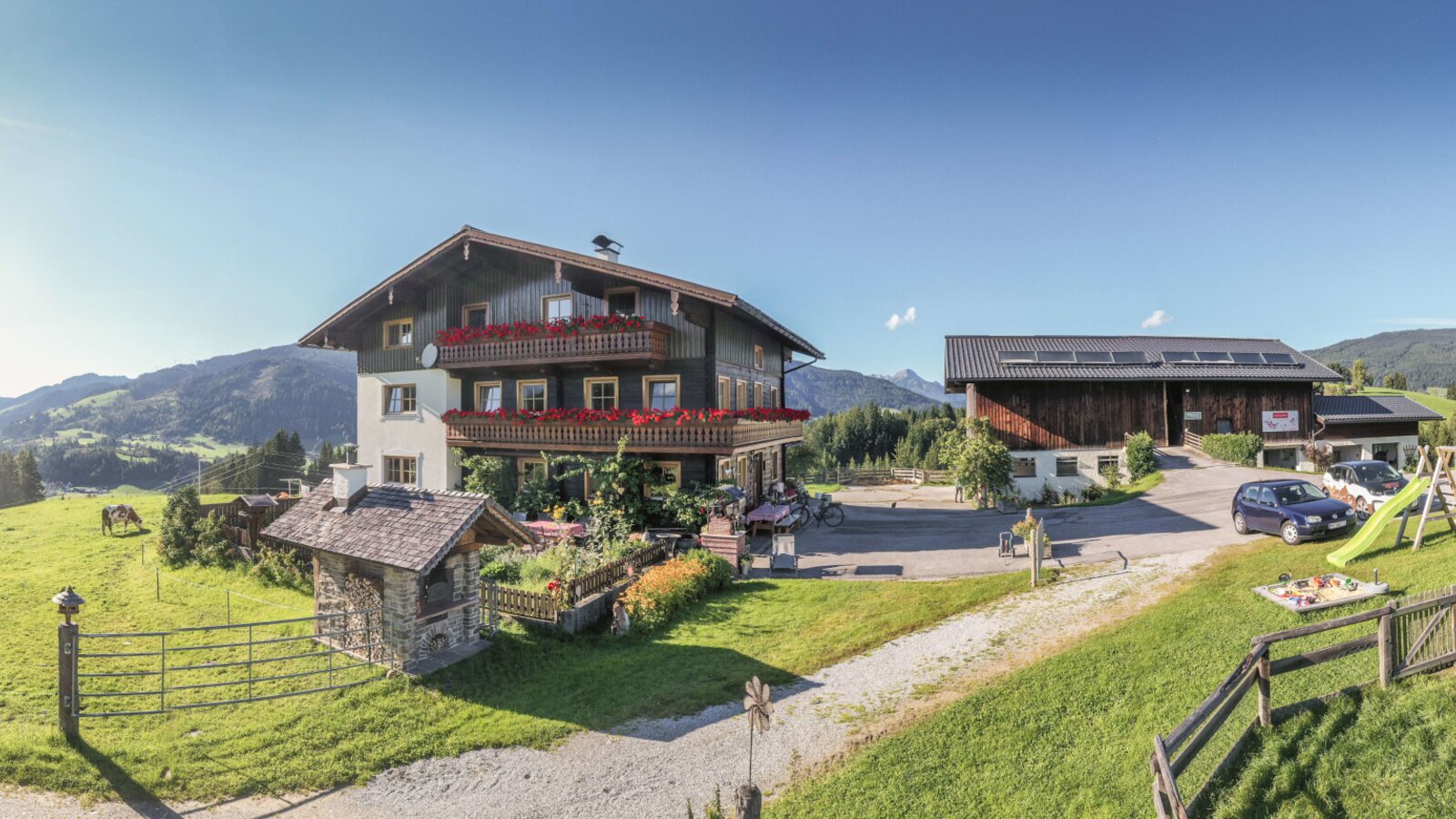 The Farm House complex includes the main building with balconies, a children's playground, and ample parking, surrounded by meadows and mountains.