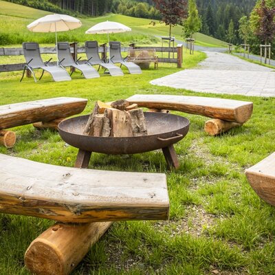 The alm's inviting outdoor area featuring a cozy fire pit surrounded by rustic wooden benches and sun loungers in the background.