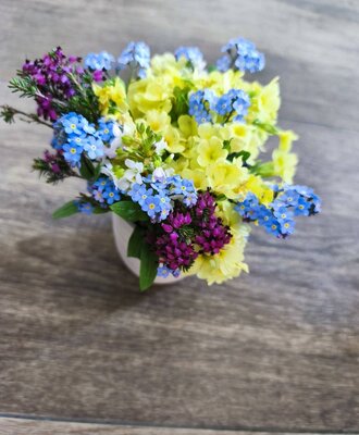 A small bouquet of yellow, blue, and purple flowers in a white vase on a wooden table at the farmhouse.