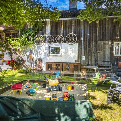 The garden area of the farm house features a sandpit with children's toys, a lawn, and sun loungers next to a traditional wooden building decorated with wagon wheels.