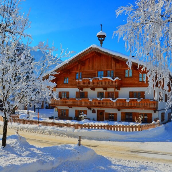 The farm house, featuring traditional wooden balconies and facade, is surrounded by a snowy winter landscape with frosted trees.