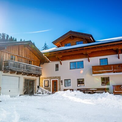 The exterior of the farm house in winter, featuring a traditional wooden facade, balconies, and snow-covered grounds with mountain views.