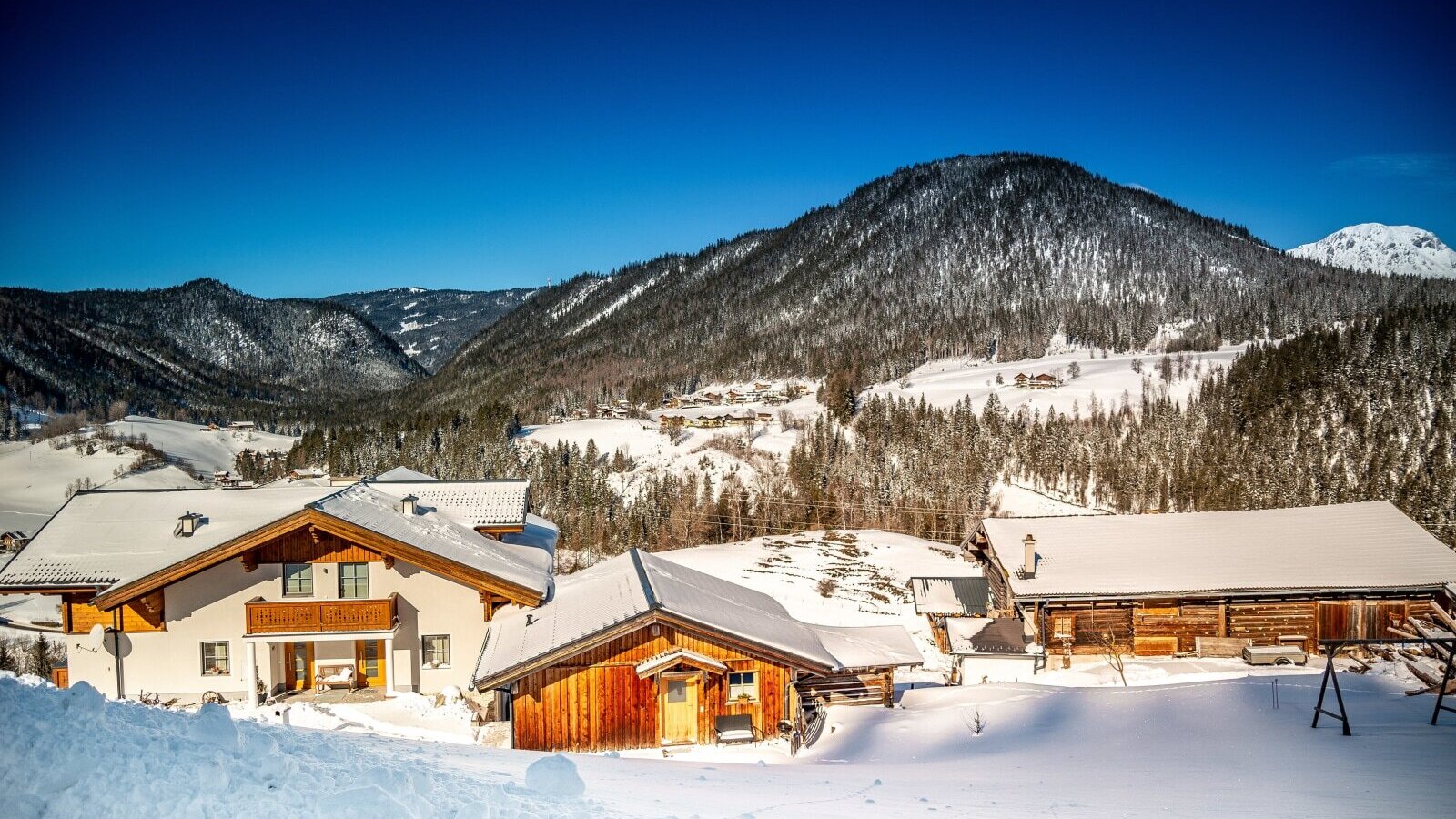 The farm house and its outbuildings are set in a snow-covered mountain landscape under a clear sky.