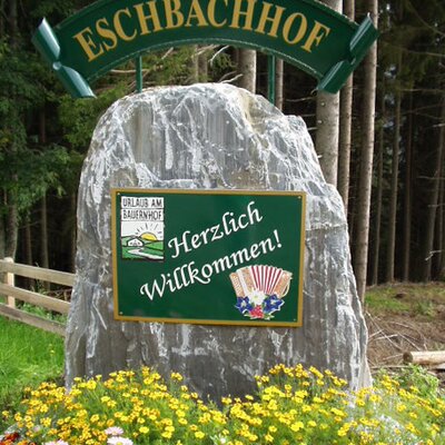 The welcome sign for the farmhouse, displaying "ESCHBACHHOF," "Herzlich Willkommen!" (Welcome!), and the "Urlaub am Bauernhof" (Holiday on the Farm) emblem, set on a large rock with colorful flowers.