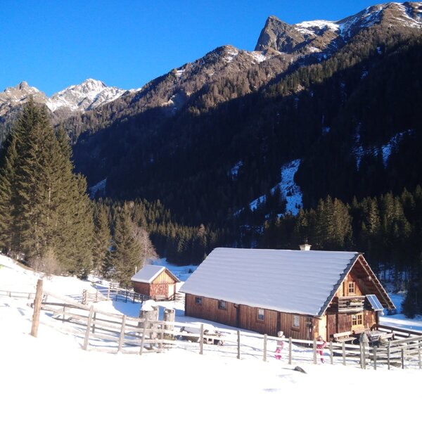 The wooden farmhouse, with a snow-covered roof and a terrace area, is set in a snowy mountain landscape with pine trees and includes a smaller adjacent outbuilding.