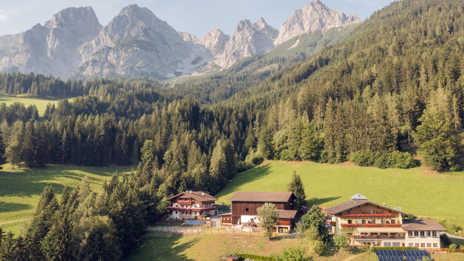 The farmhouse and its buildings are nestled in green meadows, surrounded by forests and mountains.