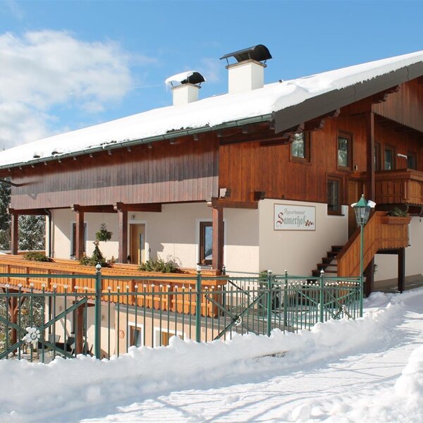 The exterior of the Farm House, named 'Apartment Samerhof' on the building, features a wooden upper level, light-colored lower level, balconies, and an outdoor staircase, situated in a snowy environment.