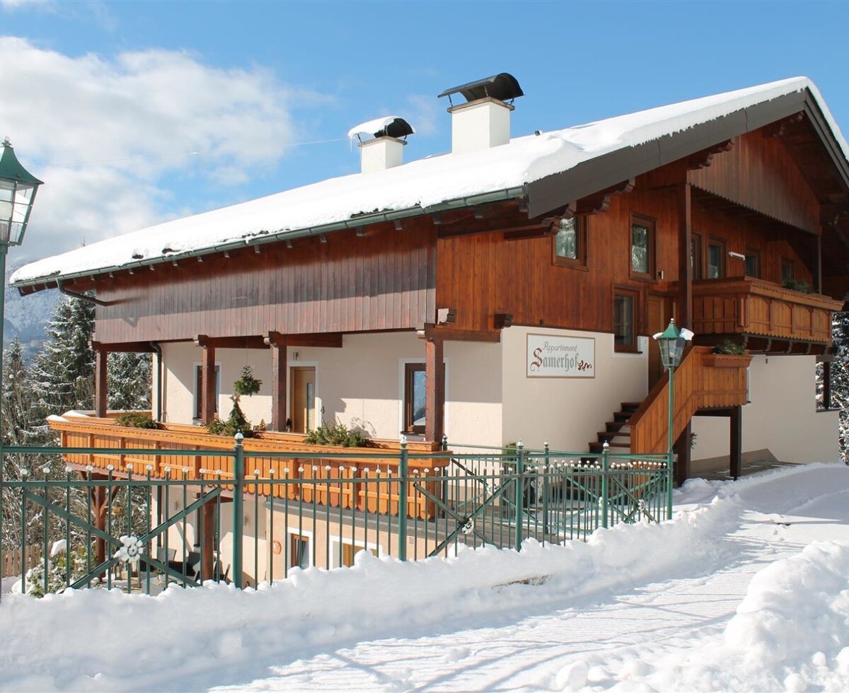 The exterior of the Farm House, named 'Apartment Samerhof' on the building, features a wooden upper level, light-colored lower level, balconies, and an outdoor staircase, situated in a snowy environment.
