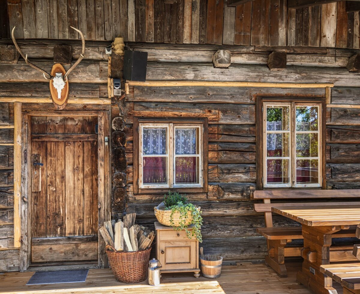 The farmhouse features a log wall exterior, a wooden entrance door with deer antlers, and an outdoor dining set on the porch.