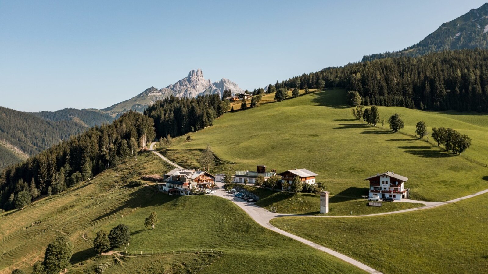 An aerial view of the Farm House complex, set on a green hillside surrounded by forests and distant mountains.