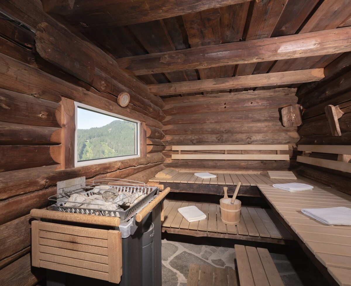 The sauna in the farmhouse with wooden beams, a sauna heater, benches, and a window offering a mountain view.