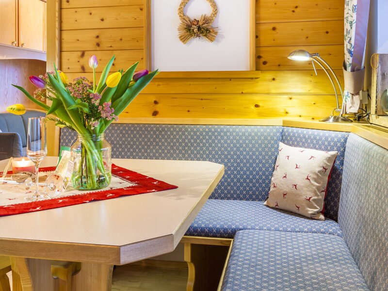 The farmhouse's dining area features a wooden table and a built-in corner bench with blue cushions, set within a wood-paneled interior.