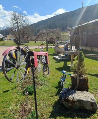The farmhouse garden with a child playing at the water trough, a decorative tractor, and mountain views.