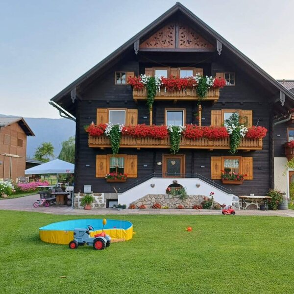 Exterior view of the farmhouse featuring flower-adorned balconies and a garden with a lawn, children's paddling pool, and toy tractor.