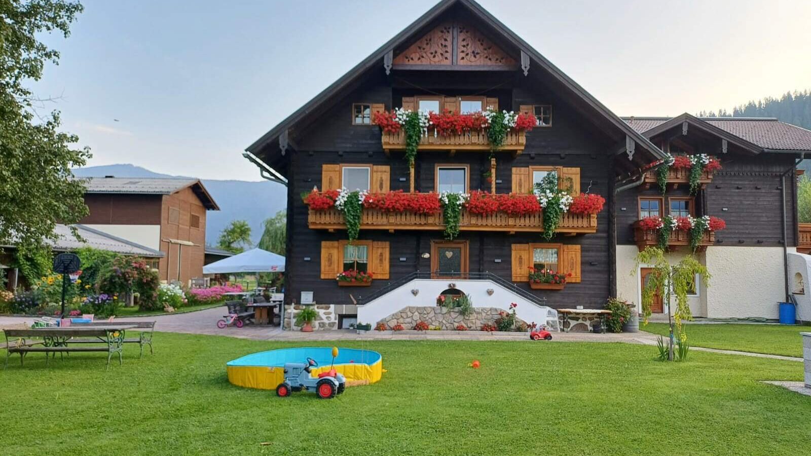 Exterior view of the farmhouse featuring flower-adorned balconies and a garden with a lawn, children's paddling pool, and toy tractor.