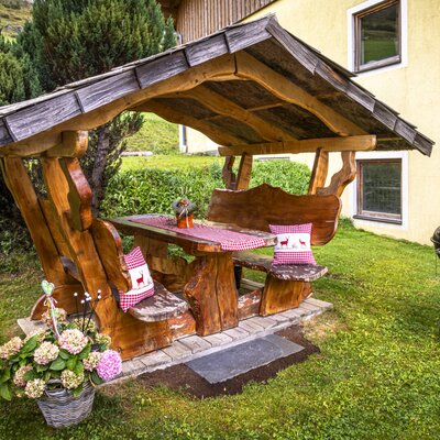 An inviting covered wooden seating area in the farmhouse's garden, featuring rustic benches with cozy cushions and an outdoor barbecue grill.