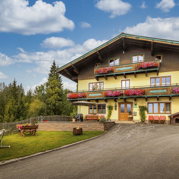 An inviting alpine farmhouse featuring flower-adorned balconies, a green garden with a hammock, and a scenic forest backdrop.