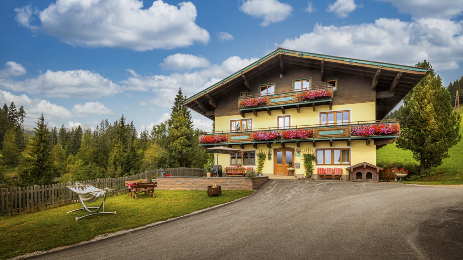 An inviting alpine farmhouse featuring flower-adorned balconies, a green garden with a hammock, and a scenic forest backdrop.