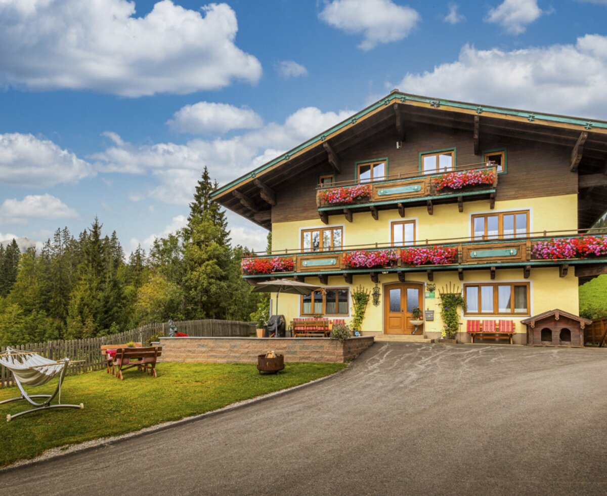 An inviting alpine farmhouse featuring flower-adorned balconies, a green garden with a hammock, and a scenic forest backdrop.