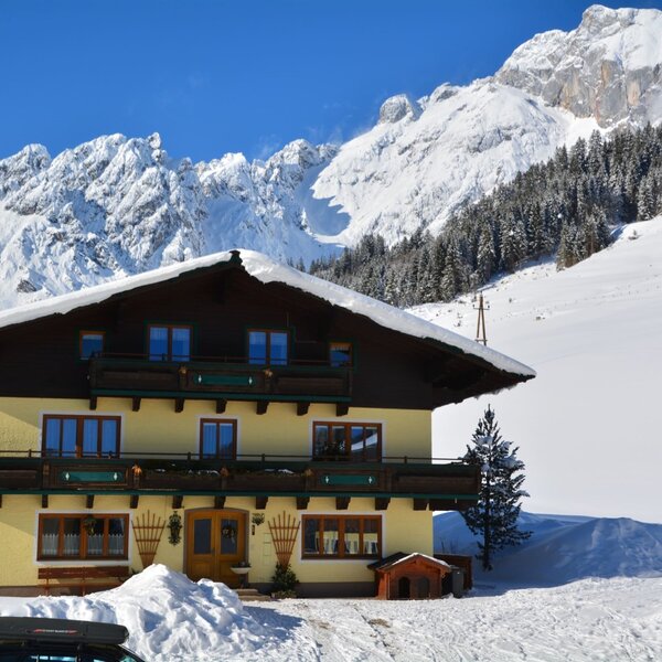 The Farm House exterior in winter, with snow-covered grounds and roof, featuring balconies and surrounded by snow-capped mountains.