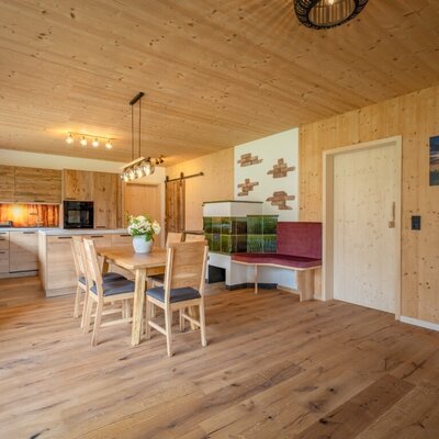 The farmhouse's open-plan kitchen and dining area features wooden cabinetry, a dining table, and a traditional tiled stove with an integrated bench.