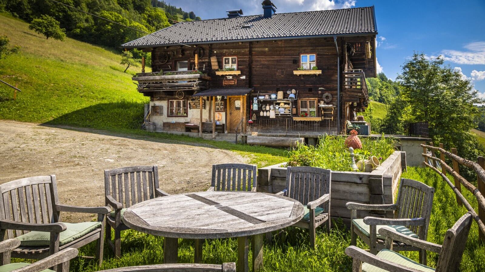 Outdoor wooden dining table and chairs with cushions on a grassy area, set in front of the traditional wooden vacation rental on a green hillside.