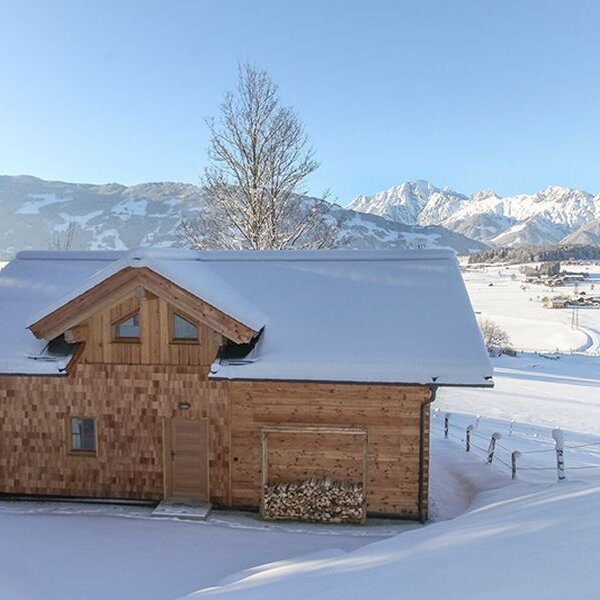 The farmhouse with its wooden exterior and stacked firewood, set in a snowy landscape with views of distant mountains.