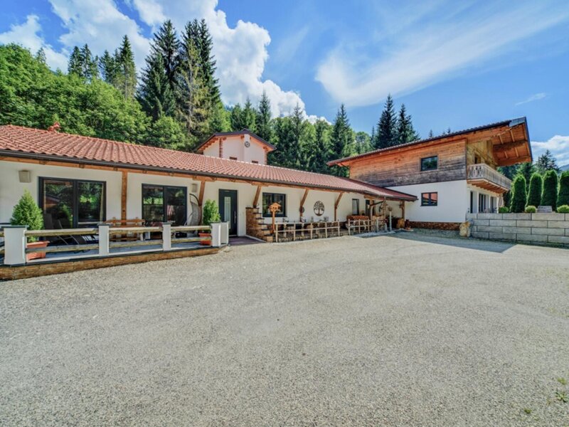 Exterior view of the holiday apartment with a large gravel parking lot and a terrace with seating, surrounded by forest and mountains.