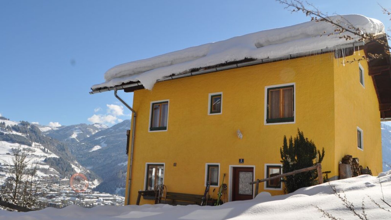 The yellow farmhouse exterior in winter, featuring snow on the roof and ground, skis by the entrance, and a view of the snowy mountains and a distant village.
