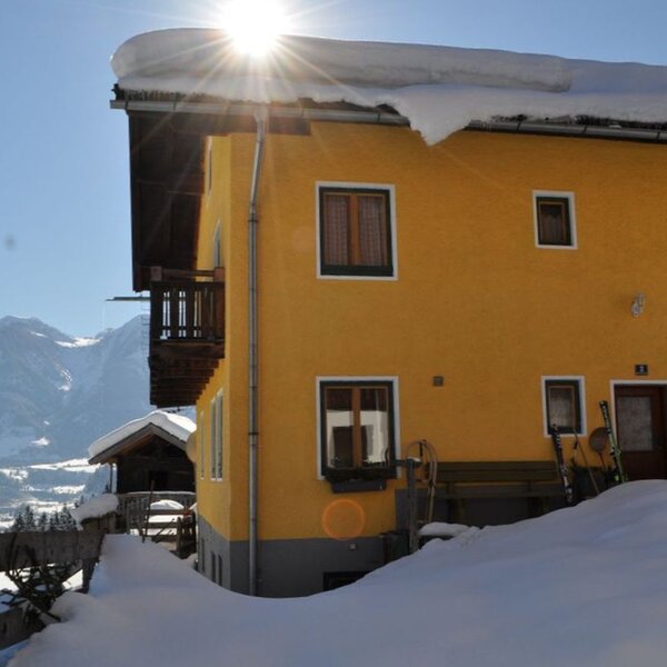 The yellow farmhouse exterior in winter, featuring a balcony, snow-covered grounds, and skis by the entrance with mountains in the background.