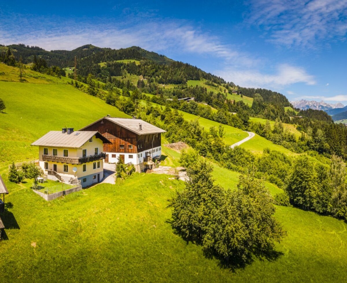 The farmhouse is situated on a green hillside with scattered trees, offering views of distant mountains.