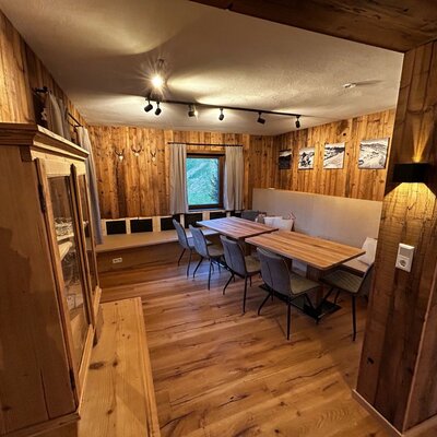 The dining area in the farmhouse features two tables with chairs, built-in bench seating, and wooden paneled walls, offering a view through the window.