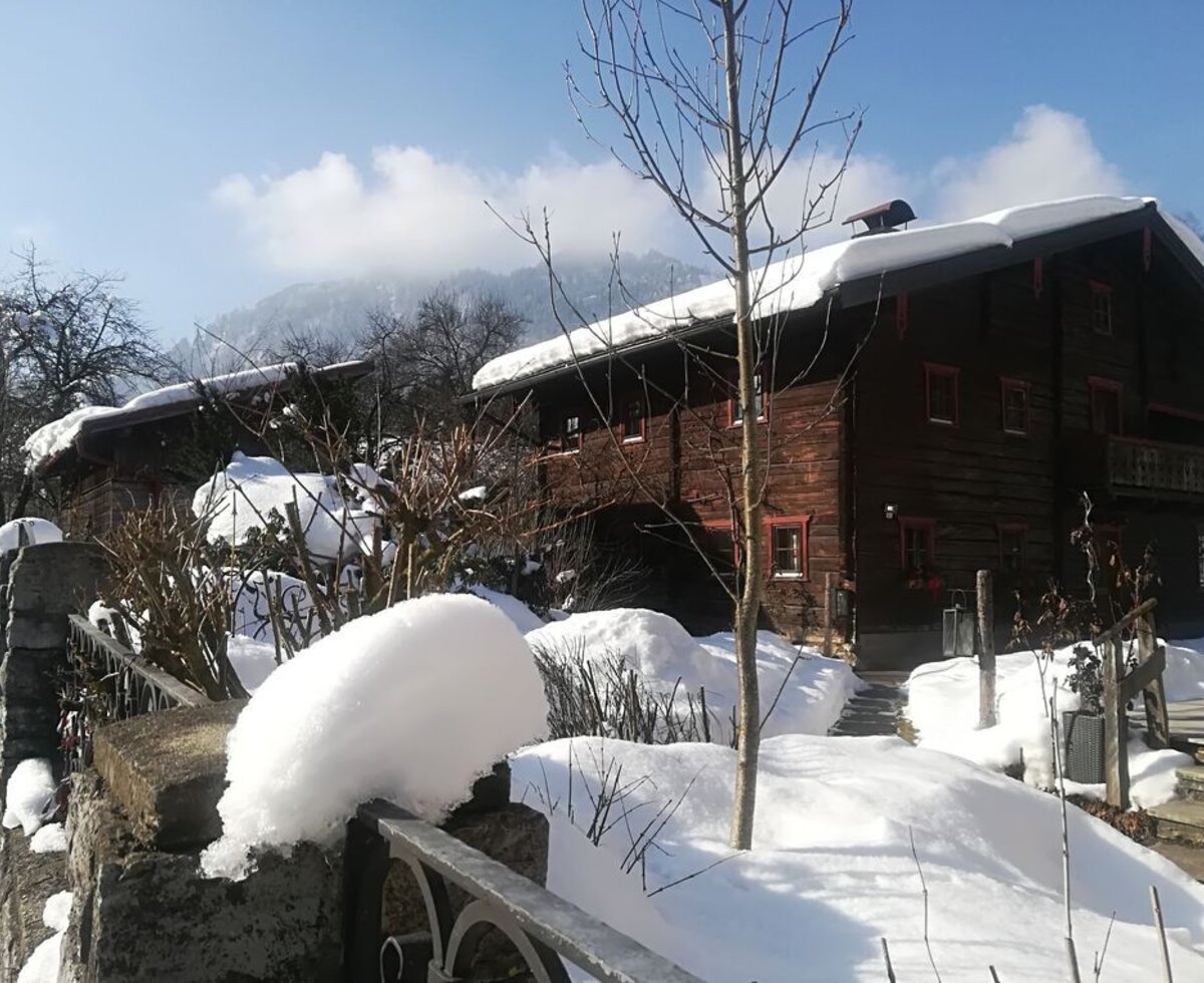 The traditional wooden farmhouse in winter, with snow covering the roof and surrounding landscape, and a path leading to the entrance.